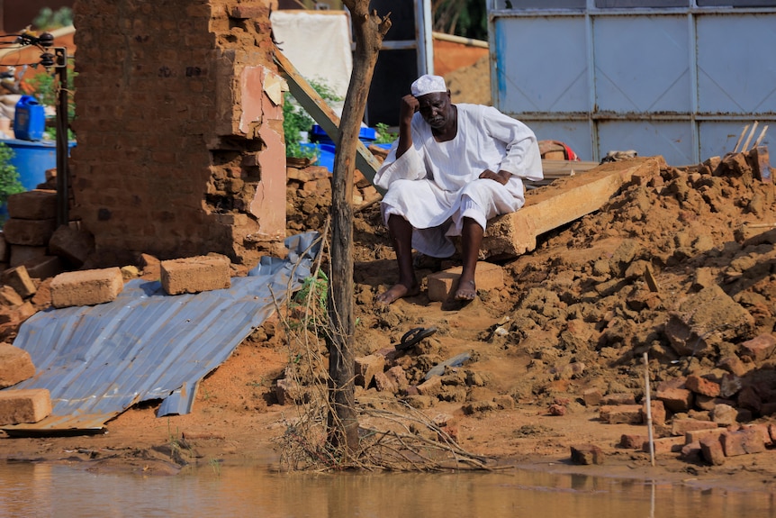 A man rests his head on his hands, sitting on the rubble of his house at the flood water's edge