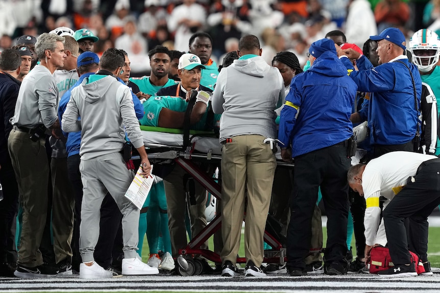 An NFL player lies on a stretcher on a field during a game as he is surrounded by coaches and medical staff.