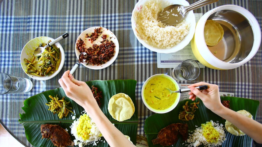 A set of Keralan dishes on white plates and banana leaf, set on a tablecloth, with a hand reaching across.