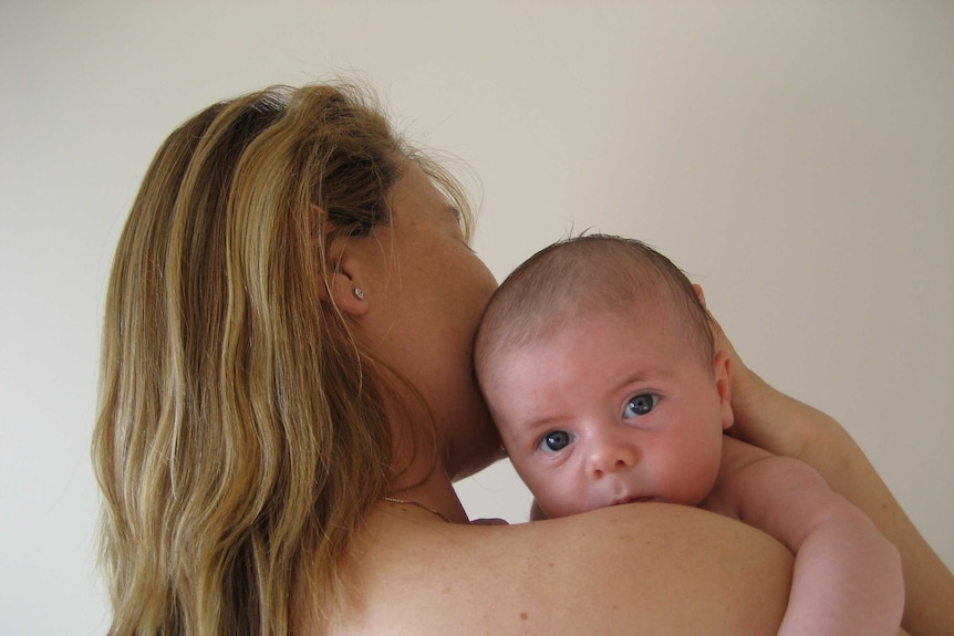 A woman with long hair looks away from the camera, holding a small baby over her shoulder.