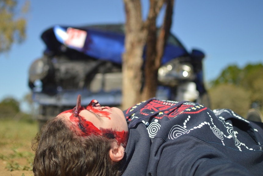 A student pretending to be dead in the foreground, with a crashed car in the background.