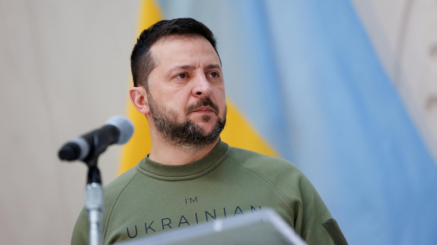 Zelenskyy in front of a Ukrainian flag wearign a khaki t shirt 
