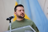 Zelenskyy in front of a Ukrainian flag wearign a khaki t shirt 
