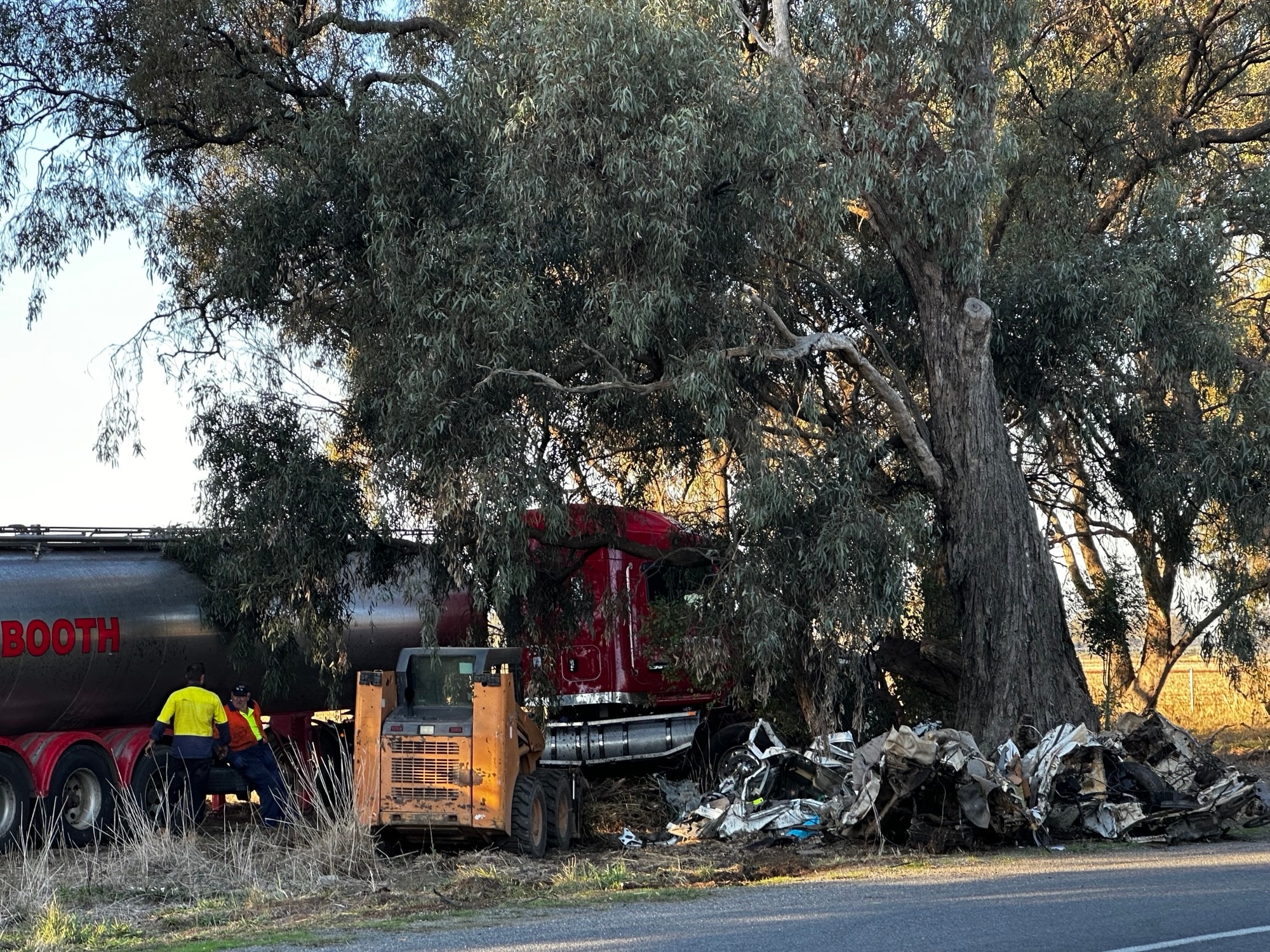 The mangled wreckage of a car which has been damaged beyond recognition against a tree, with a large truck behind it.