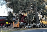 The mangled wreckage of a car which has been damaged beyond recognition against a tree, with a large truck behind it.