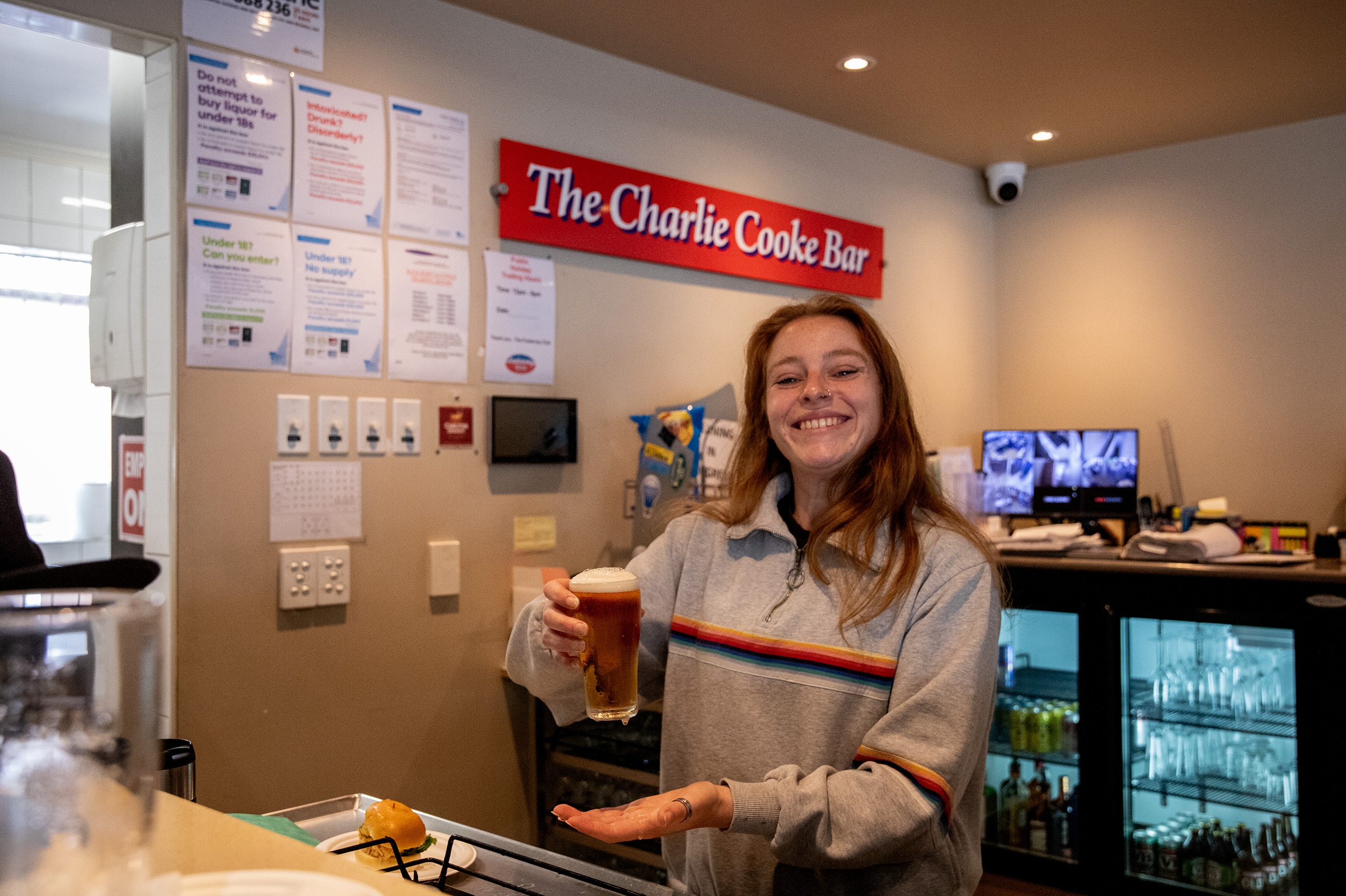 A woman stands behind a bar holding a glass of beer. She is smiling, has long hair past her shoulders. There is a fridge behind.