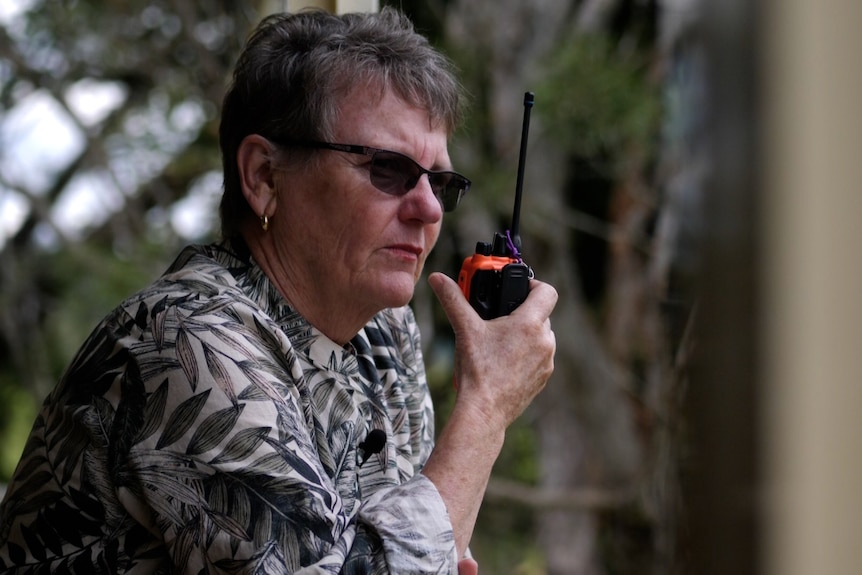 A woman with short grey hair and glasses on a deck holding a small radio near her mouth