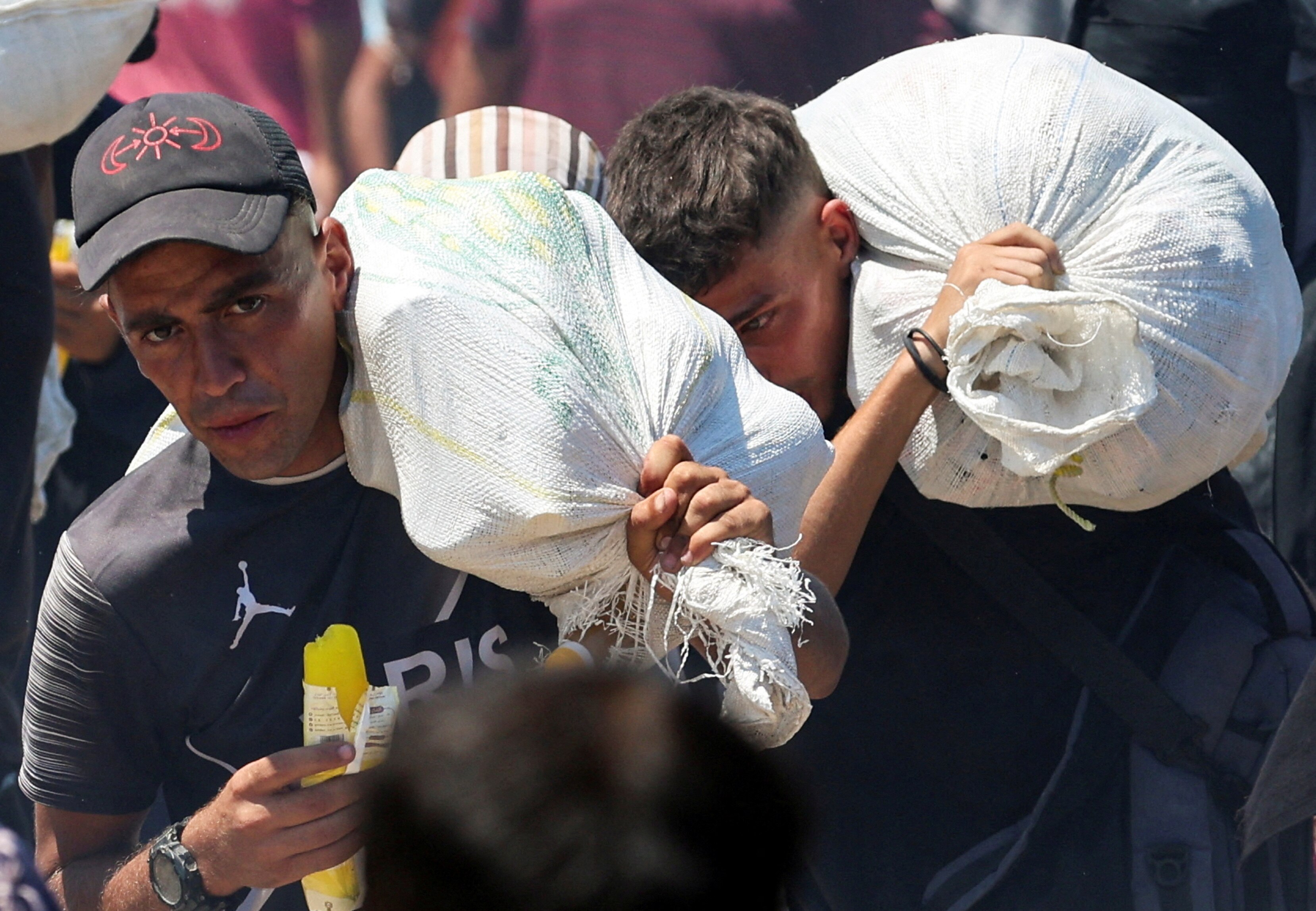 Two men carrying sacks of food in a crowd.