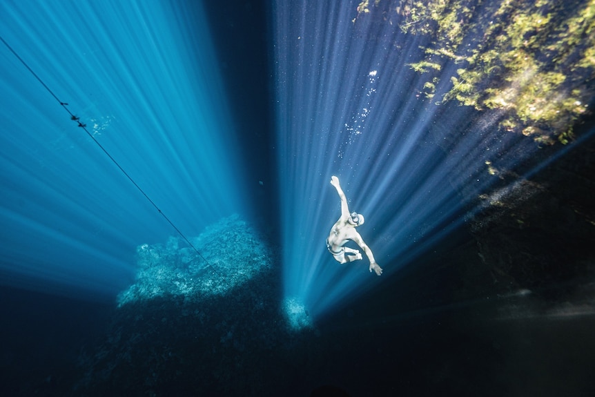 A diver in a wetsuit wearing a snorkel floats up to the surface under a bright light beam.
