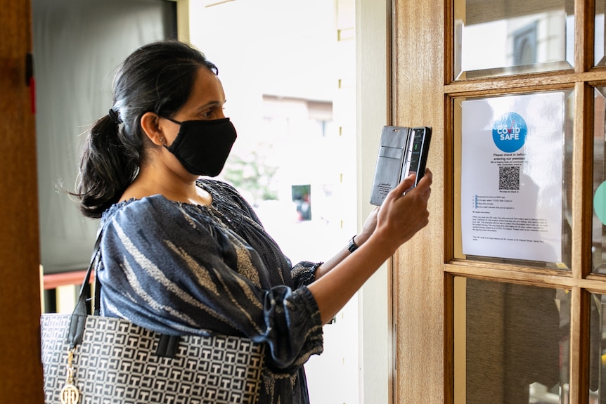 A woman wearing a face mask holds up  her mobile phone to a QR code. 