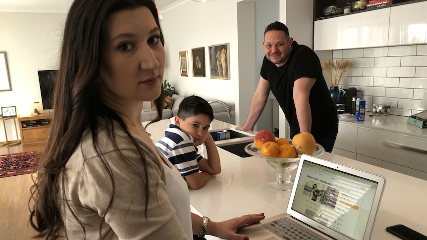 A woman, man and young boy in an open-plan kitchen. The woman has a GoFundMe page open on a laptop sitting on the kitchen bench