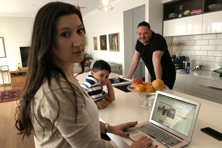 A woman, man and young boy in an open-plan kitchen. The woman has a GoFundMe page open on a laptop sitting on the kitchen bench
