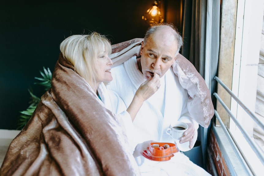 man and woman in robes sit at window under blanket eating chocolates and coffee