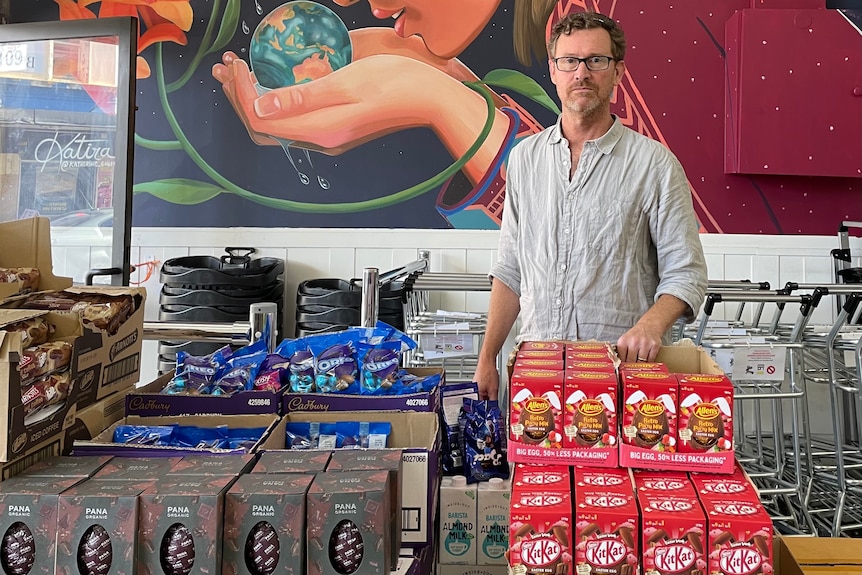 A man with glasses standing in a shop with stacks of Easter eggs.