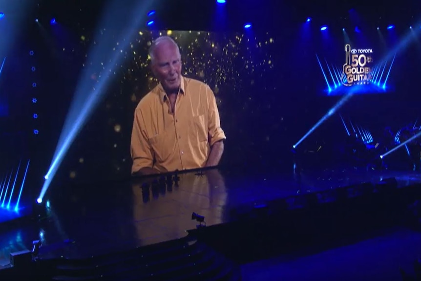 A large, low-lit stage with a large video screen showing an older man.