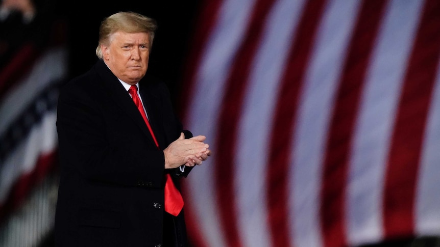 A man with blonde hair and wearing a black suit with a red tie claps his hands in front of the American flag