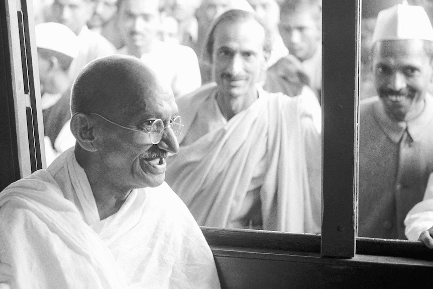 Mahatma Gandhi sits in a train carriage with his palm out, while tens of men look through the window, smiling.