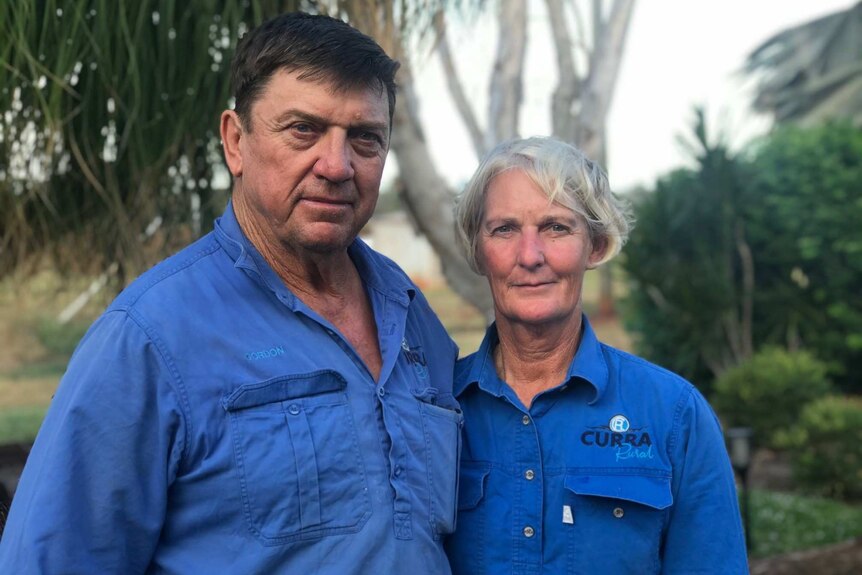 a man and woman stand looking into the camera. They're wearing their cattle farming shirts and  shrubs are in the background
