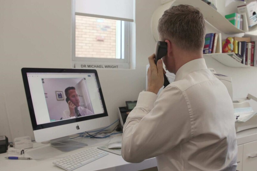 A GP sits at his office desk, holding a phone to his ear with a video call on the computer screen in front of him.