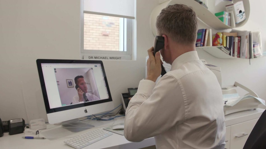 A GP sits at his office desk, holding a phone to his ear with a video call on the computer screen in front of him.