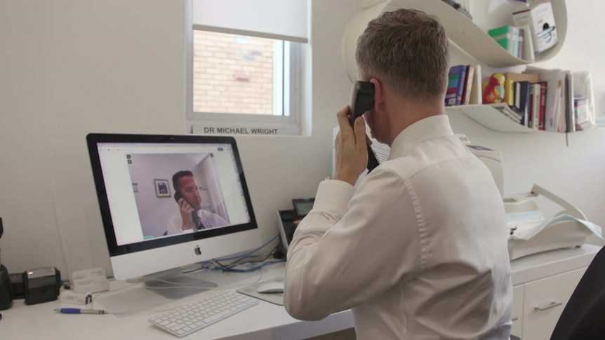 GP Michael Wright sits at his office desk, holding a phone to his ear with a video call on the computer screen in front of him.