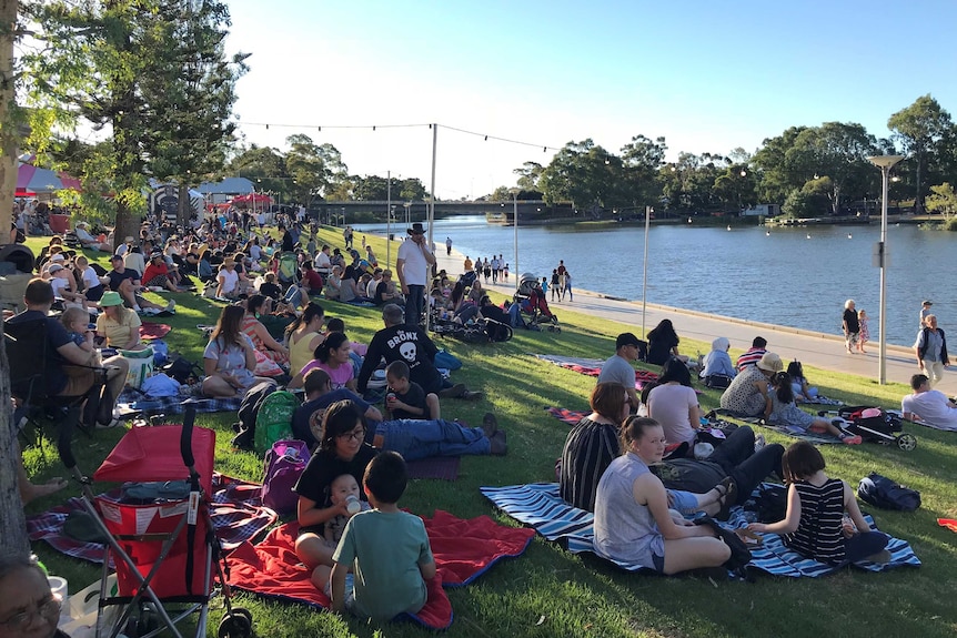 Around 100 people sit on picnic blankets looking out over Torrens Lake on a sunny New Year's Eve.