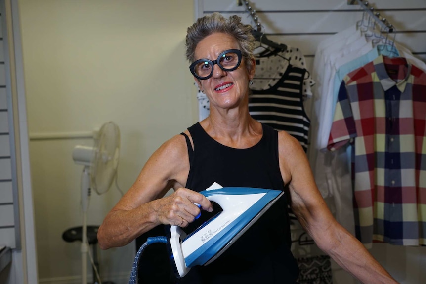 A woman irons a shirt beside a fan.