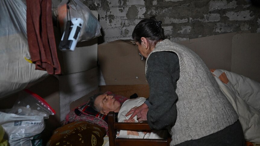 An old woman tends to an old man lying in bed, in a dark room with brick walls.