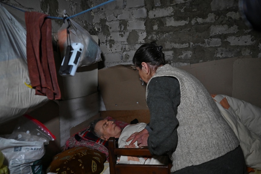 An old woman tends to an old man lying in bed, in a dark room with brick walls.