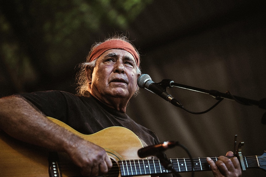 Close-up of musician Kev Carmody wearing red head band, looking into distance while on stage playing guitar and singing.