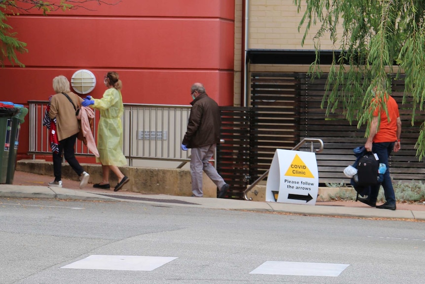 An older woman is escorted into Joondalup Private Hospital by a medical worker in a plastic gown as two men follow.