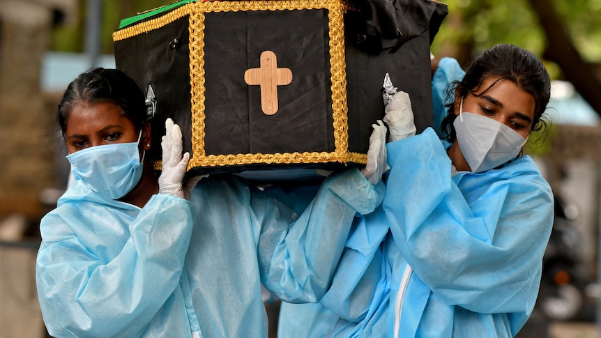 Two women wearing PPE carry a coffin with a crucifix painted on the end