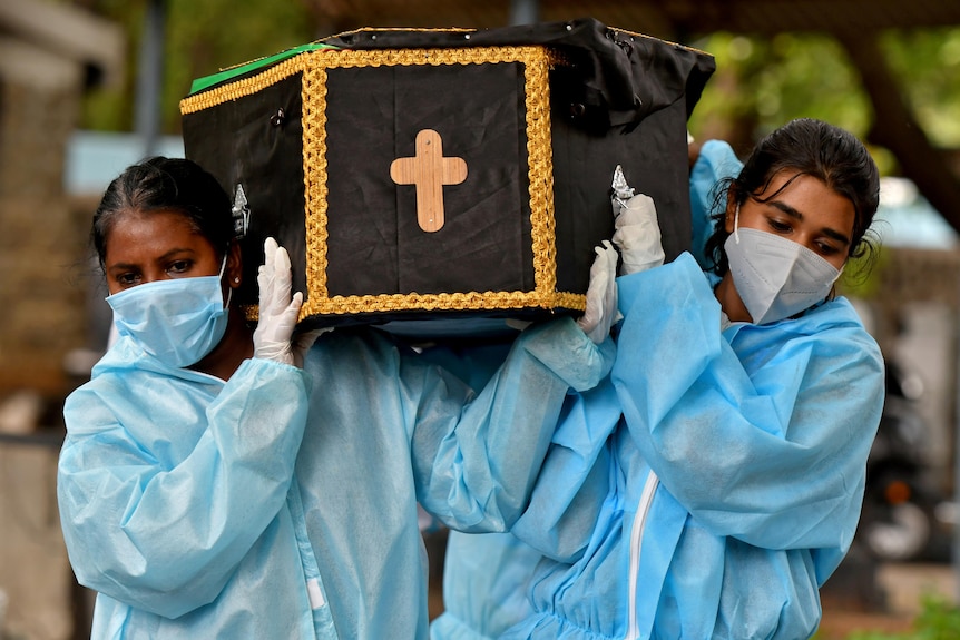 Two women wearing PPE carry a coffin with a crucifix painted on the end