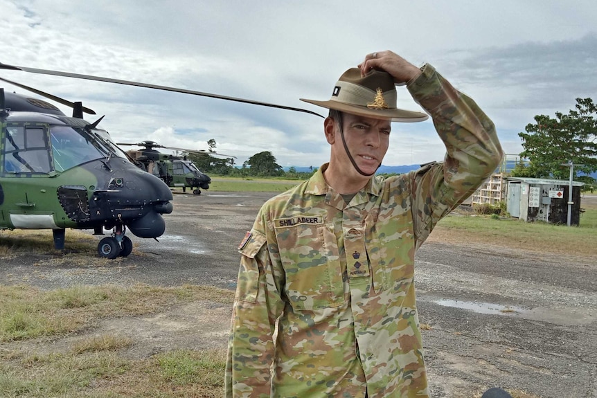Australian Defence Force Lieutenant Colonel Corey Shillabeer wears army uniform and holds onto his army hat with helicopter.