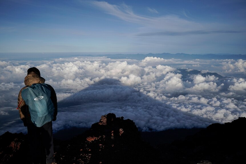 A climber wearing a green backpack looks at a mountain that has cast its shadow on clouds below