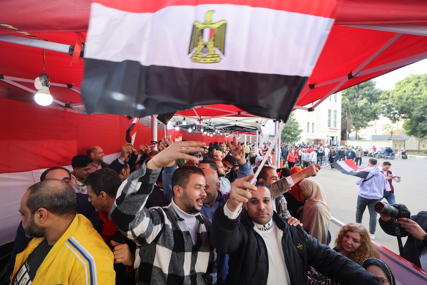 A crowd of people, some waving flags, lined up under a big red marquee