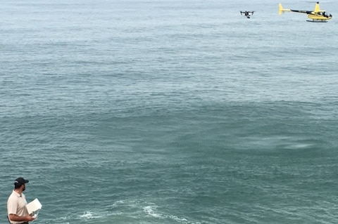Researcher on coastal headland watches drone and helicopter fly over the ocean