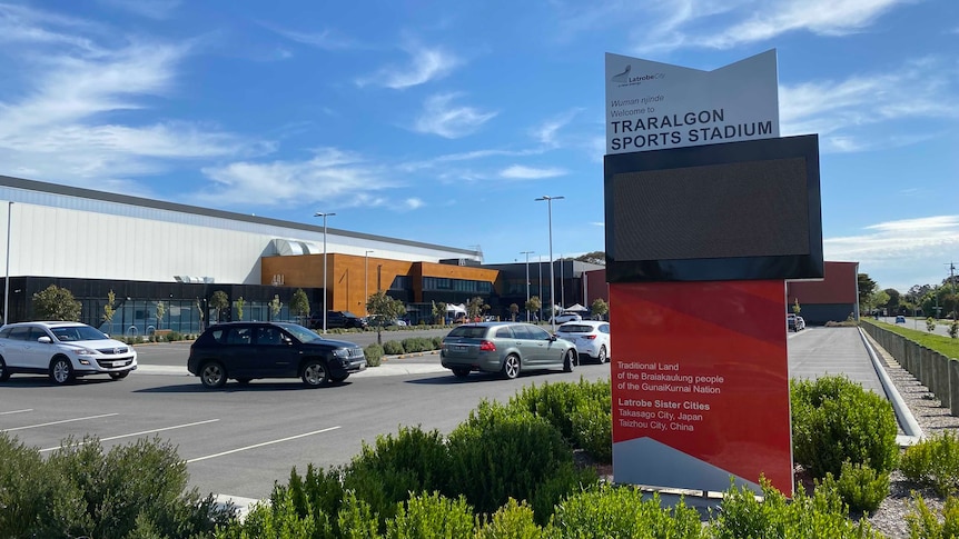 A sign for the stadium in front of a line of cars waiting for testing