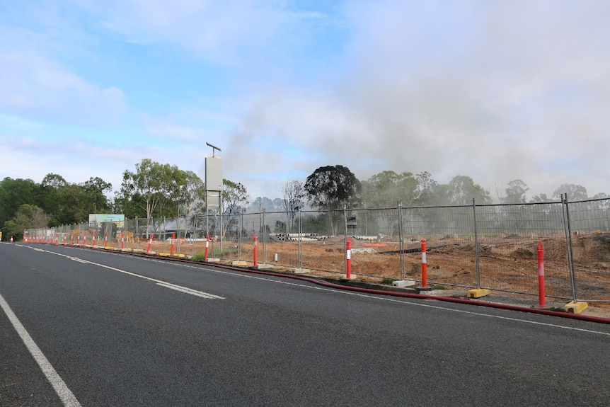 Grey smoke in the air behind a fence, with a road in the foreground.