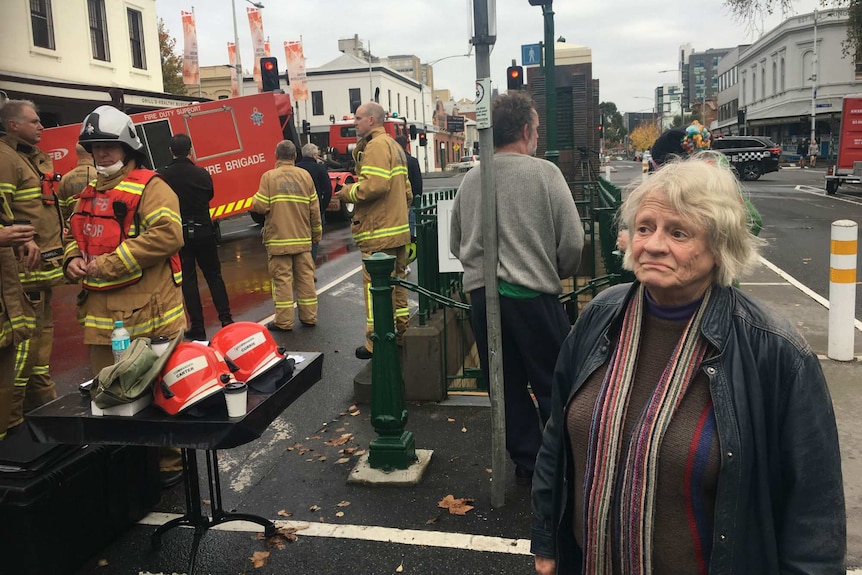 A woman stands on the street with firefighters in the background.