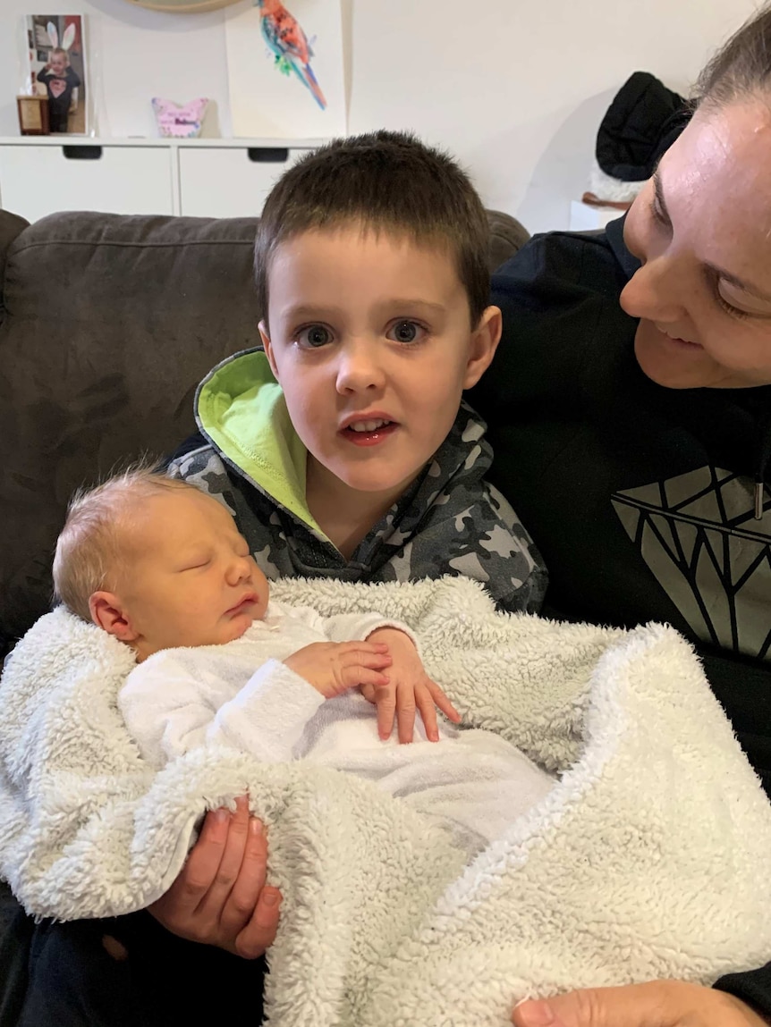 A little boy holds his newborn brother while his mum looks on.