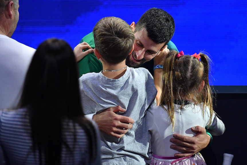 A smiling Novak Djokovic bends down to hug his children after winning a big tennis tournament.