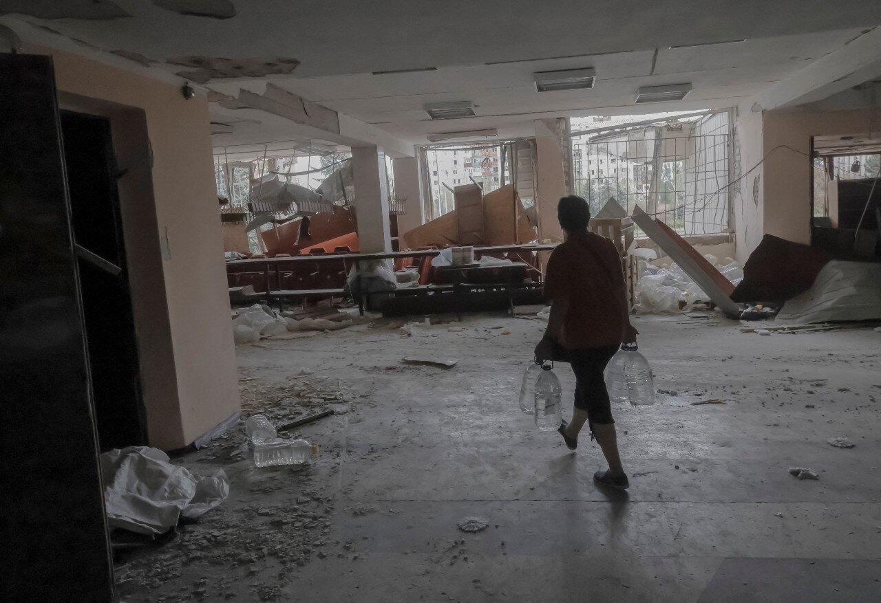 A woman carries large bottles of water through a damaged building.