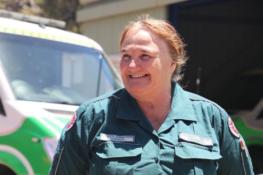 A woman wearing a green ambulance uniform smiles and stands in front of an ambulance vehicle.