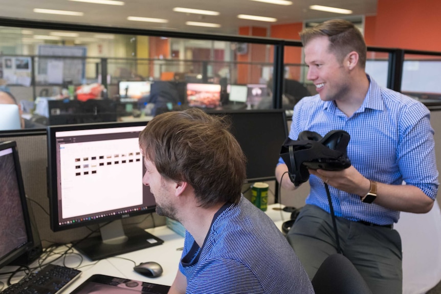 Nathan Bazley holding VR headset watching a member of the production team at work on a computer in the ABC offices.