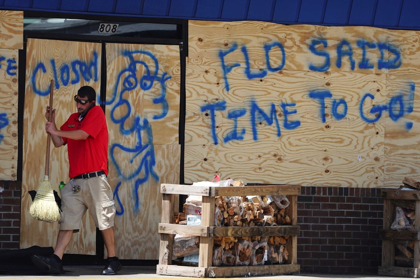 A man in an orange shirt holding a broom in front of a boarded up shop with graffiti saying 'Flo said it's time to go'