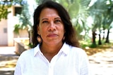A woman looks at the camera from close range during the day wearing a white shirt.