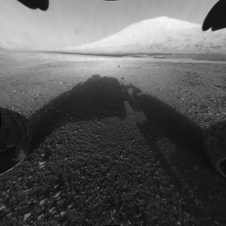The surface of Mars in black and white, showing a tall, wide Mt Sharp in the distance and bare rocks and soil in the foreground.