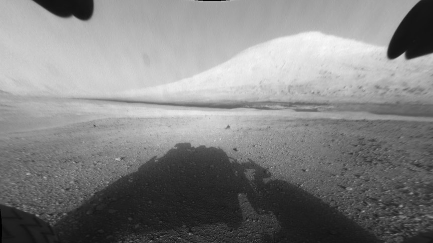 The surface of Mars in black and white, showing a tall, wide Mt Sharp in the distance and bare rocks and soil in the foreground.