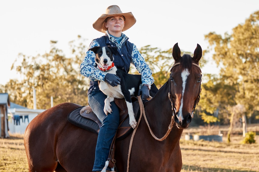 A woman sits on a horse, a dog on her lap.